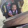 baby, child, infant, smiling, car_seat, stroller, harness, outdoor, portrait, face, brown_hair, pink_shirt, butterflies, sunlight, pavement, fabric, safety_straps, cup, yellow_liquid, happy