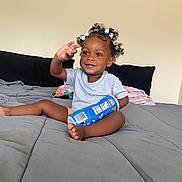 Ariella joined the competition — help win amazing prizes! toddler, child, curly_hair, beads, smiling, waving, sitting, bed, blanket, pillow, snack_container, indoor, casual_clothing, happy, person, baby, cute, room, relaxed, playful
