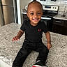 toddler, child, smiling, sitting, kitchen, countertop, black_clothing, jeans, sneakers, modern_kitchen, appliances, refrigerator, oven, microwave, happy, portrait, indoor, person, cute, young_child