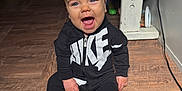 Kash is registered to the contest to win money with this photo: baby, child, smiling, indoors, floor, wooden_floor, clothing, nike, black_outfit, sitting, happy, person, face, blue_eyes, table, toy, electric_cord, wood, foot, open_mouth