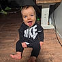 baby, child, smiling, indoors, floor, wooden_floor, clothing, nike, black_outfit, sitting, happy, person, face, blue_eyes, table, toy, electric_cord, wood, foot, open_mouth
