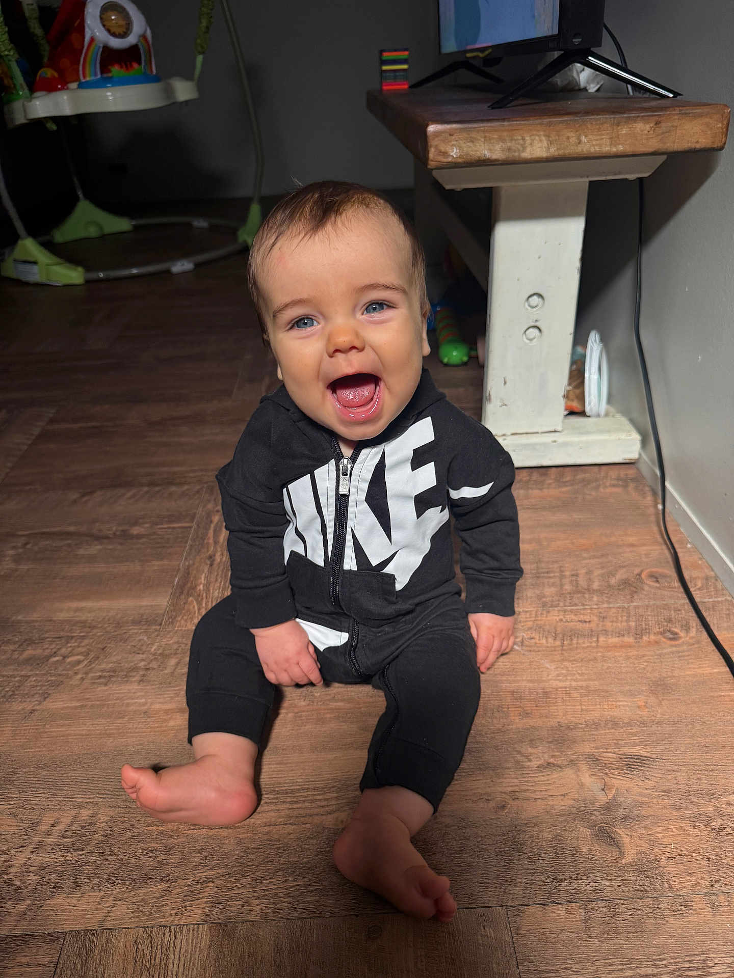 Kash is registered to the contest to win money with this photo: baby, child, smiling, indoors, floor, wooden_floor, clothing, nike, black_outfit, sitting, happy, person, face, blue_eyes, table, toy, electric_cord, wood, foot, open_mouth