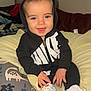 Kash is registered to the contest to win money with this photo: baby, child, hoodie, black_clothing, white_shoes, smiling, blue_eyes, blanket, pillow, dinosaur_pattern, indoors, seated, young_child, cute, happy, casual_clothing, footwear, portrait, infant, cozy