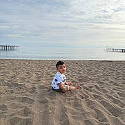 Mikael joined the competition — help win amazing prizes! toddler, child, beach, sand, sea, sky, cloudy, water, pier, shore, outdoor, nature, summer, casual_clothing, shorts, tshirt, barefoot, sitting, curious, daytime