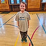 Tyler Jr. is registered to the contest to win money with this photo: child, boy, smiling, tshirt, gymnasium, indoor, wooden_floor, camouflage_pants, green_shoes, sports_lines, standing, casual_clothing, short_hair, happy, young, portrait, playful, big_brother, person, background_windows