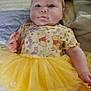 Olivia joined the competition — help win amazing prizes! baby, child, tutu, yellow_dress, cartoon_print, bed, blanket, cushion, soft, cute, infant, lying_down, chubby_cheeks, smile, patterned_clothing, indoors, person, young_child, face, expression