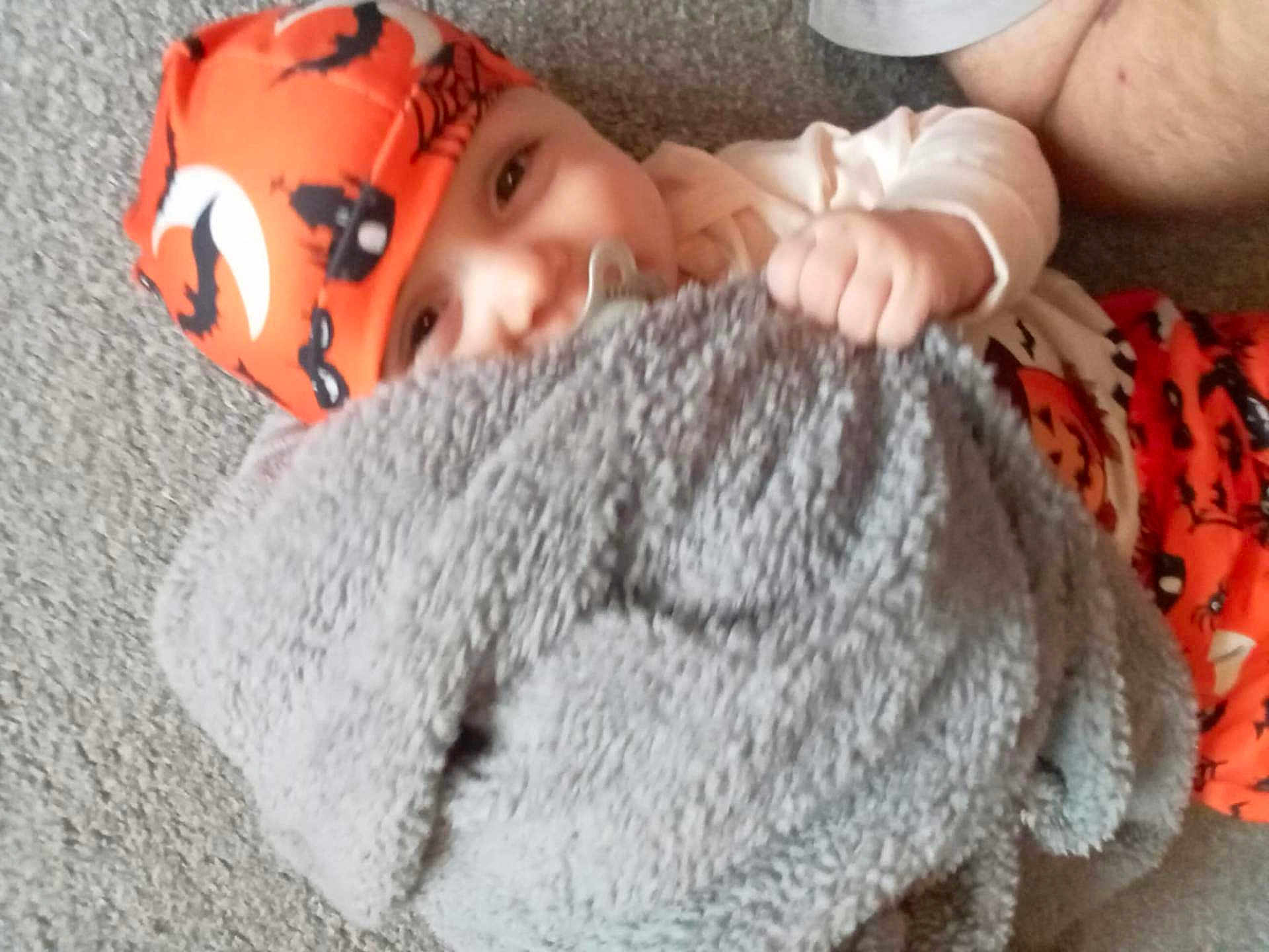 Noah joined the competition — help win amazing prizes! baby, child, pacifier, blanket, orange_hat, halloween_theme, carpet, floor, smiling, cute, infant, clothing, indoor, playful, hand, headwear, costume, pumpkin_pattern, person, face