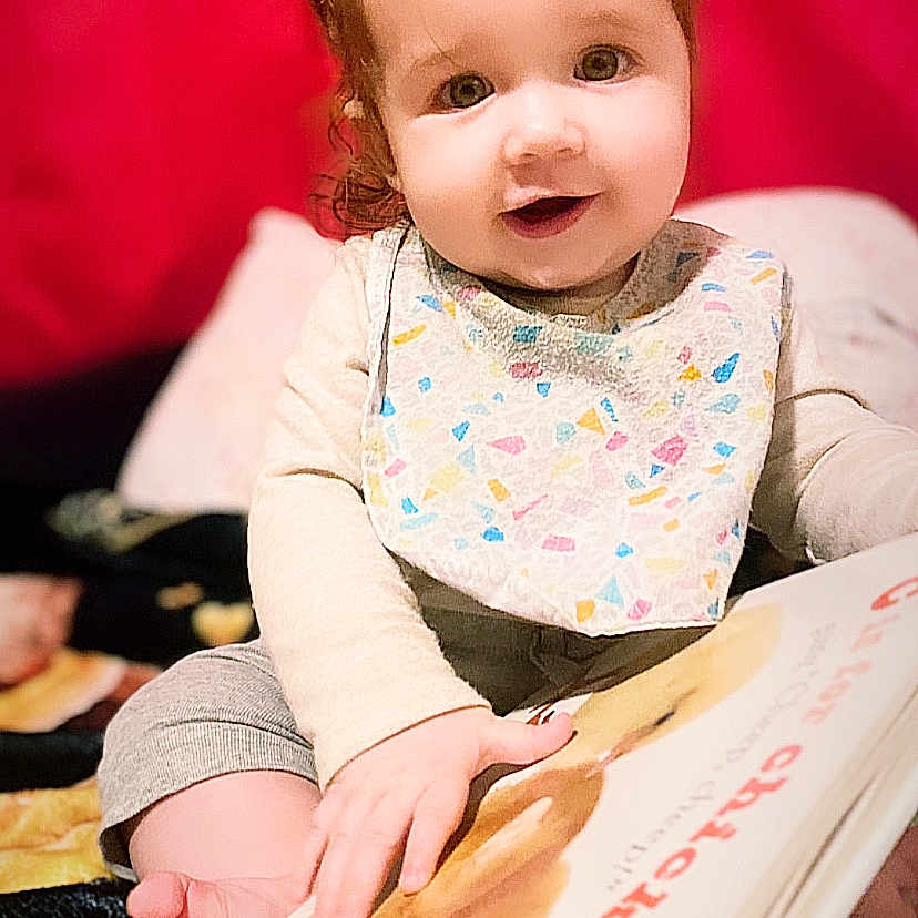 Alora is registered to the contest to win money with this photo: applique, baby, bodypart, clothing, cutlery, dress, face, finger, hand, happy, head, pattern, person, photography, plywood, portrait, reading, sitting, smile, wood