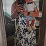 woman, baby, mirror, selfie, dress, floral_pattern, indoor, person, holding, baby_clothes, bow_tie, suspenders, footwear, carpet, window, curtains, bed, television, smartwatch, reflection