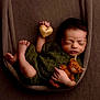 newborn, baby, sleeping, teddy_bear, heart, green_outfit, brown_background, fabric, cozy, cute, infant, portrait, resting, child, soft_lighting, wrapped, peaceful, tiny_feet, handheld_toy, closeup