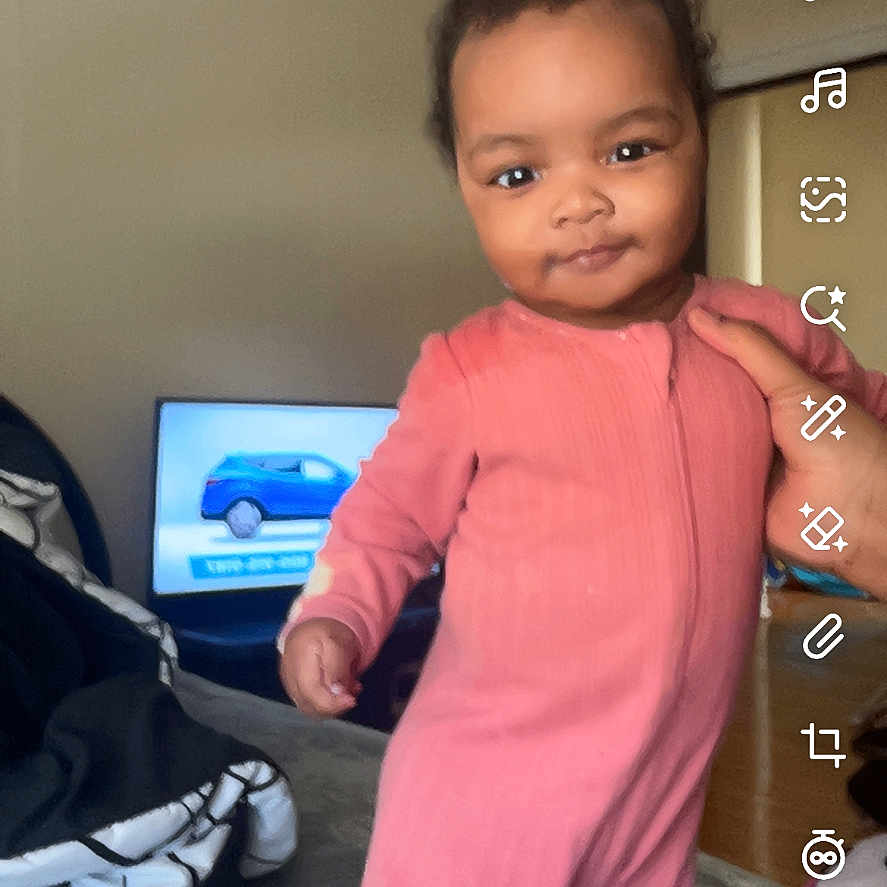 Aasheeda is registered to the contest to win money with this photo: baby, blanket, blue_car, casual, child, cozy, curly_hair, floor, furniture, hand, home, indoor, person, pink_onesie, room, smile, standing, television, toddler, wall