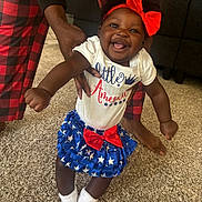 Paislee is registered to the contest to win money with this photo: baby, child, smiling, indoor, carpet, red_bow, blue_skirt, white_socks, hands, adult_support, person, happy, cute, patriotic, clothing, footwear, home, family, celebration, fun