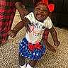 baby, child, smiling, indoor, carpet, red_bow, blue_skirt, white_socks, hands, adult_support, person, happy, cute, patriotic, clothing, footwear, home, family, celebration, fun