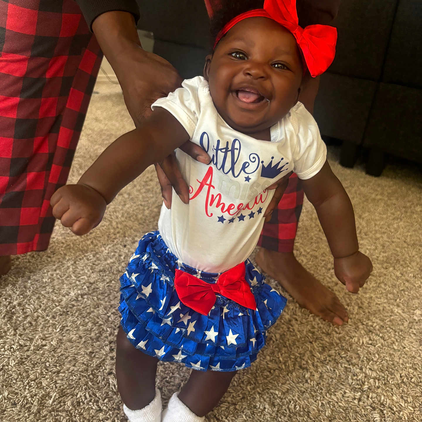 Paislee is registered to the contest to win money with this photo: baby, child, smiling, indoor, carpet, red_bow, blue_skirt, white_socks, hands, adult_support, person, happy, cute, patriotic, clothing, footwear, home, family, celebration, fun