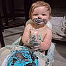 baby, child, high_chair, cake, blue_frosting, messy, eating, smiling, portrait, indoor, kitchen, chair, food, dessert, birthday, cream, hands, face, blond_hair, happy