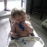 Wyatt is registered to the contest to win money with this photo: baby, child, high_chair, spoon, mealtime, food, plate, kitchen, indoor, sitting, blond_hair, face, serious_expression, blue_spoon, tray, adult, slippers, floor, sunlight, home