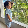 toddler, child, standing, portrait, window, indoor, tropical_garden, palm_leaves, plants, greenery, sunlight, blue_sky, curly_hair, blue_outfit, barefoot, looking_at_camera, home, wooden_floor, glass_window, daylight