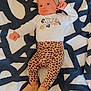 baby, infant, headband, baby_clothes, patterned_pants, footwear, blanket, abstract_pattern, lying_down, smiling, cute, face, person, child, indoors, cozy, fashion, apparel, portrait, soft_lighting