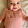 baby, infant, smile, face, onesie, pink_clothing, text_on_clothing, lying_down, blanket, cute, happy, portrait, child, person, closeup, young_child, skin, hand, soft_texture, apparel