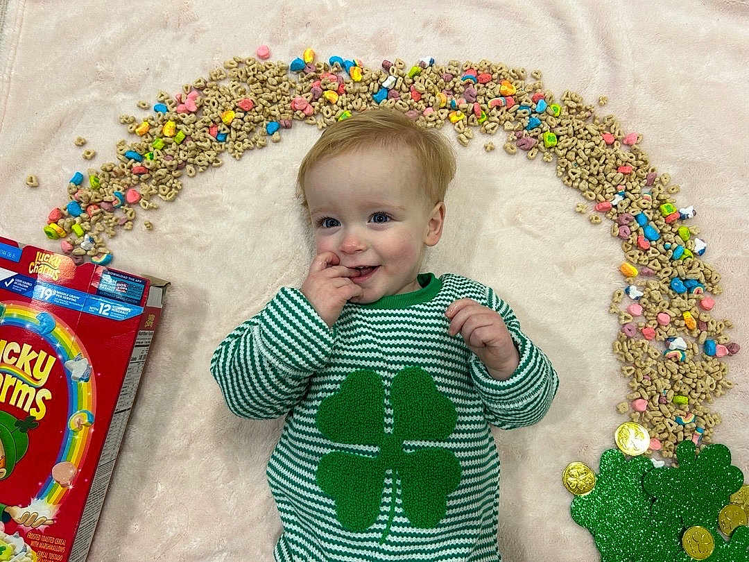 Theodore joined the competition — help win amazing prizes! child, baby, infant, smile, green_sweater, four_leaf_clover, cereal, lucky_charms_box, marshmallows, blanket, laying_down, portrait, happy, hand_to_mouth, gold_coins, colorful, toy, striped_clothing, indoor, cute