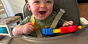 Theodore is registered to the contest to win money with this photo: baby, blue_eyes, child, clothing, face, furniture, green, happy, high_chair, indoor, kitchen, person, plastic_toy, red, seat_belt, smiling, table, toy, yellow, young_child