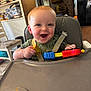 baby, high_chair, toy, smiling, happy, indoor, child, face, blue_eyes, clothing, kitchen, furniture, plastic_toy, seat_belt, red, yellow, green, person, young_child, table