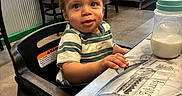 Zakai joined the competition — help win amazing prizes! airplane_design, baby_bottle, casual_clothing, child, concrete_floor, curious, curly_hair, furniture, high_chair, indoor, metal_wall, milk, person, plastic_chair, restaurant, seated, striped_shirt, table, toddler, wood_paneling