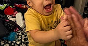 Zakai is registered to the contest to win money with this photo: toddler, child, yellow_shirt, clapping, hands, blanket, bed, indoor, television, adult_hand, joyful, expression, hair, face, room, door, picture_frame, furniture, person, home
