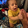 toddler, child, yellow_shirt, clapping, hands, blanket, bed, indoor, television, adult_hand, joyful, expression, hair, face, room, door, picture_frame, furniture, person, home