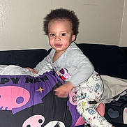 Zakai is registered to the contest to win money with this photo: toddler, child, cushion, skull_pattern, blanket, sofa, indoor, wall_art, floral_picture, pajamas, footwear, smiling, face, hair, clothing, home, colorful, playful, portrait, person