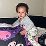 toddler, child, cushion, skull_pattern, blanket, sofa, indoor, wall_art, floral_picture, pajamas, footwear, smiling, face, hair, clothing, home, colorful, playful, portrait, person