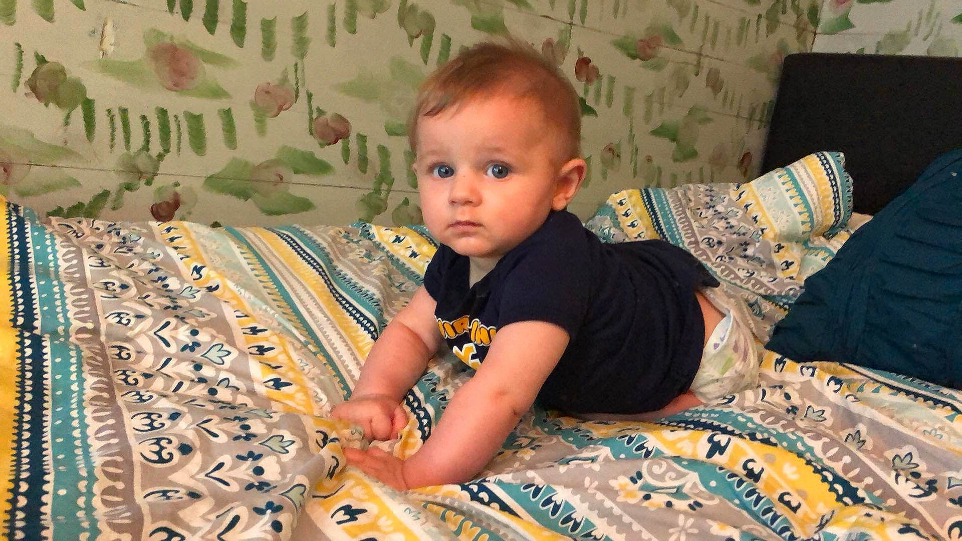 Ezra is registered to the contest to win money with this photo: baby, blanket, child, linens, person, quilt, room, sitting, smile, textile, toddler