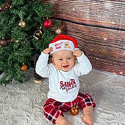 Ezio a rejoint le concours — aidez-le/la à gagner de superbes lots ! baby, child, christmas_tree, santa_hat, plaid_pants, white_rug, holiday, festive, decorations, ornaments, smiling, indoor, wooden_background, red, white, toddler, sitting, cute, happy, celebration