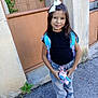 child, girl, backpack, plush_toy, white_bow, jeans, black_shirt, sneakers, outdoor, pavement, gate, wall, plant, smile, standing, casual_clothing, daylight, young, happy, portrait