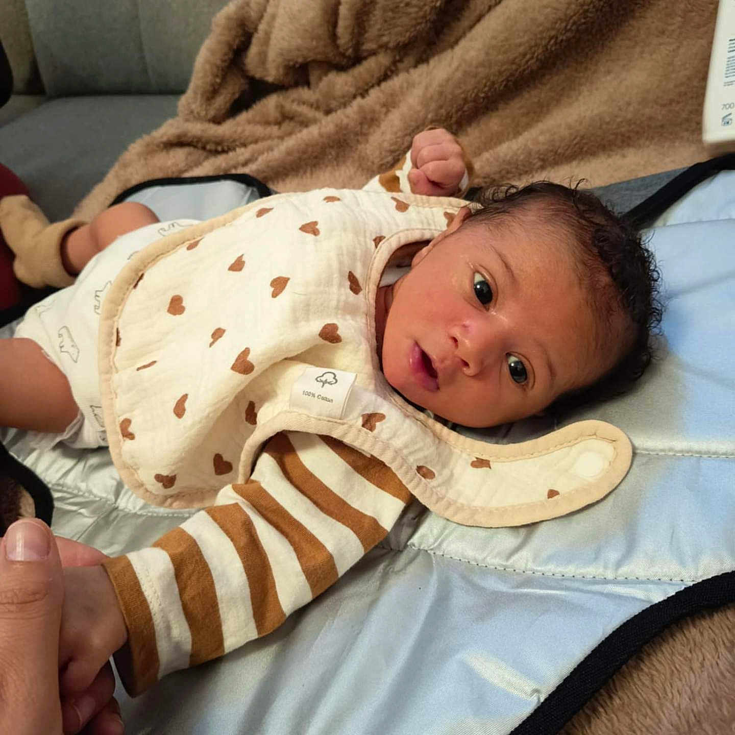 Jahyan a rejoint le concours — aidez-le/la à gagner de superbes lots ! baby, bib, blanket, brown_heart_pattern, cozy, curly_hair, cute, expression, face, hand, holding_hand, indoor, infant, lying_down, newborn, person, skin, soft_texture, striped_clothing, tender_moment