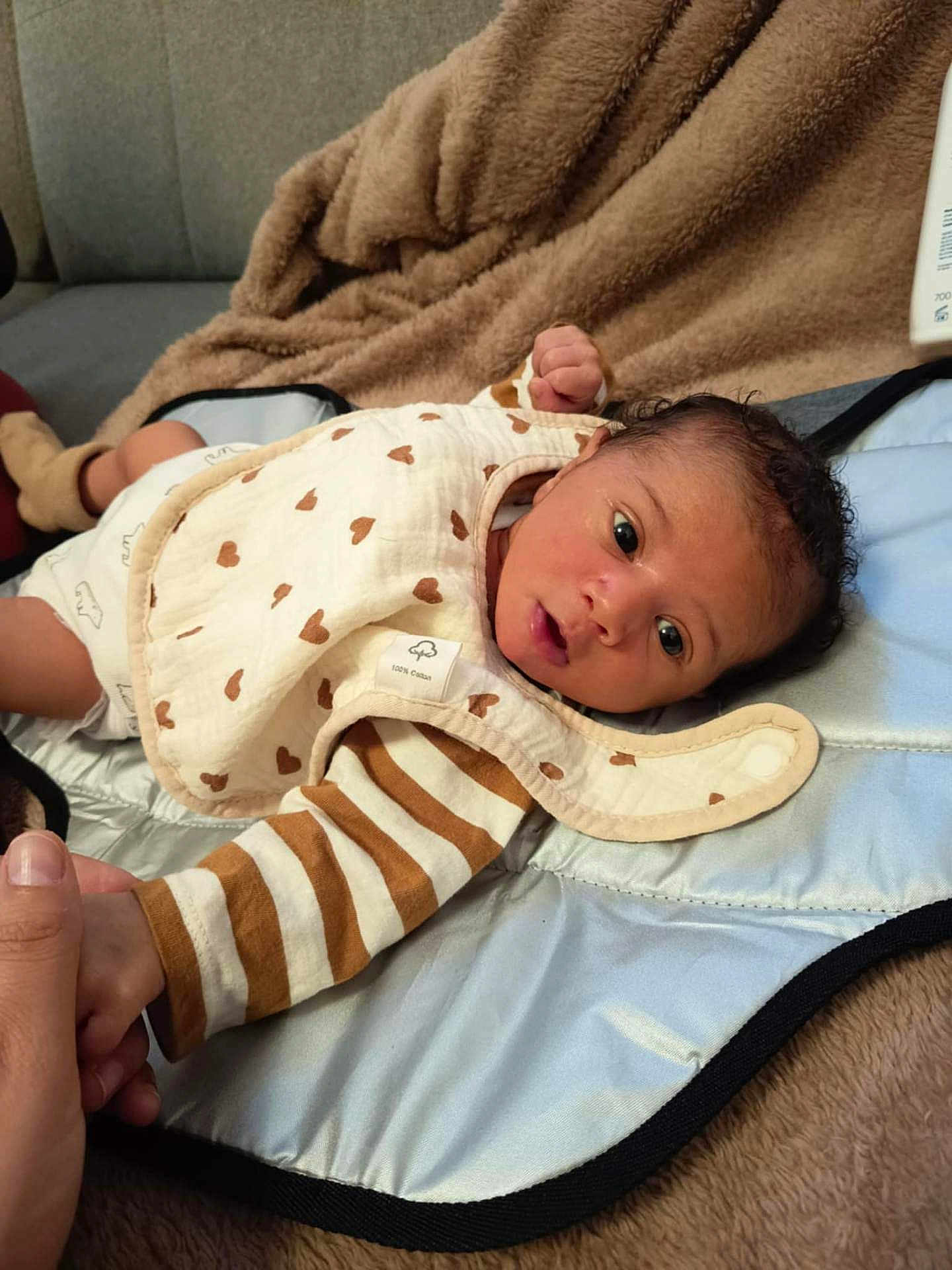 Jahyan a rejoint le concours — aidez-le/la à gagner de superbes lots ! baby, infant, face, hand, blanket, bib, striped_clothing, brown_heart_pattern, curly_hair, lying_down, soft_texture, indoor, person, skin, cute, newborn, holding_hand, cozy, expression, tender_moment