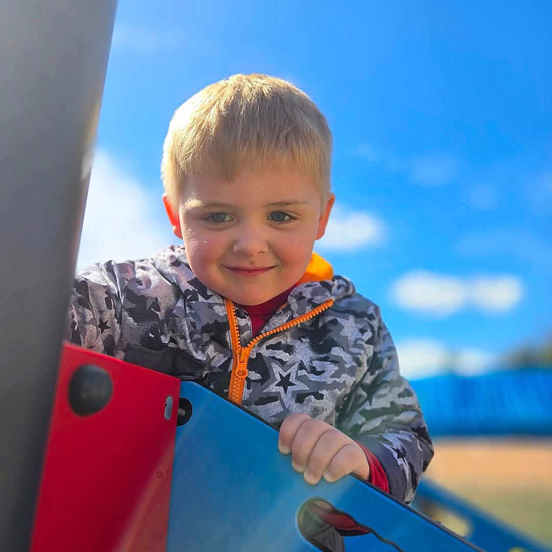 Kane is registered to the contest to win money with this photo: blonde_hair, blue_sky, boy, camouflage, casual_clothing, child, climbing, daylight, fun, hand, happy, jacket, nature, outdoor, person, play_equipment, playground, recreation, smile, young