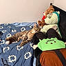 Fro is registered to the contest to win money with this photo: cat, tabby_cat, bed, blue_bedspread, stuffed_animals, teddy_bear, dinosaur_toy, plush_toy, indoor, feline, relaxed, lying_down, curious, goggles, bedroom, cozy, soft_toys, patterned_bedspread, resting, pet
