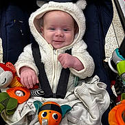 Jamie is registered to the contest to win money with this photo: baby, infant, stroller, hooded_jacket, white_clothing, smile, toy, plush_toy, car_seat, child, cute, indoor, shopping_cart, blanket, person, face, happy, small_hands, seatbelt, wearing