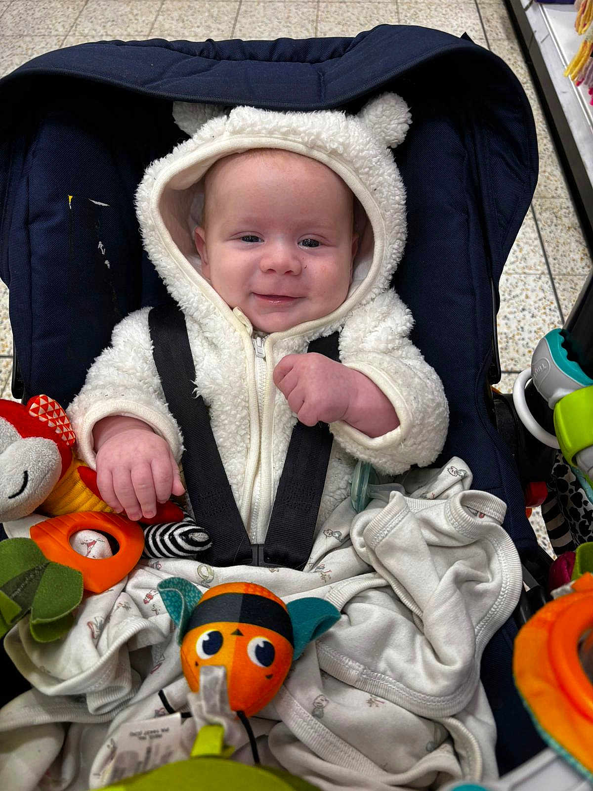Jamie is registered to the contest to win money with this photo: baby, infant, stroller, hooded_jacket, white_clothing, smile, toy, plush_toy, car_seat, child, cute, indoor, shopping_cart, blanket, person, face, happy, small_hands, seatbelt, wearing