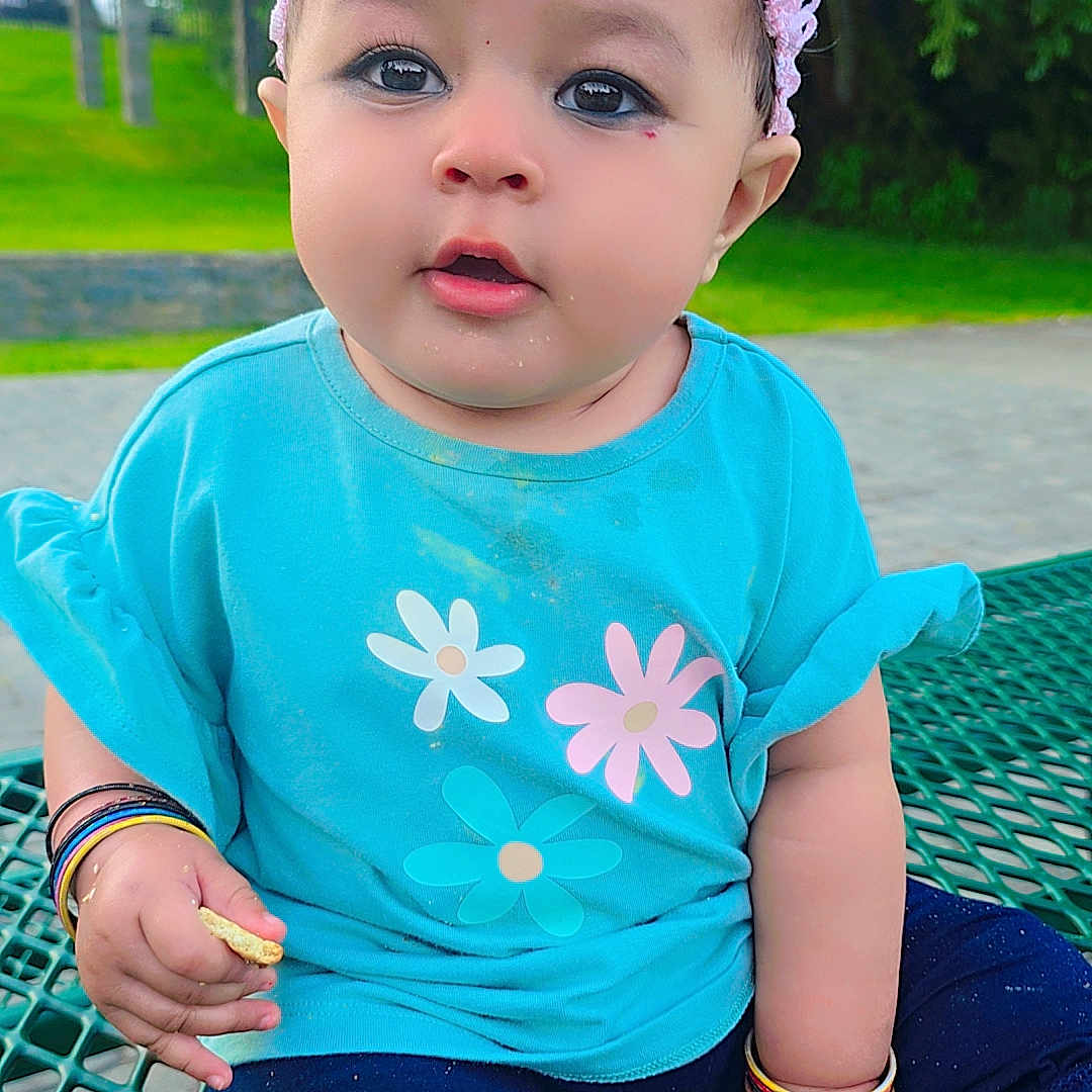 Miral is registered to the contest to win money with this photo: baby, toddler, child, outdoor, picnic_table, headband, flower, bracelets, blue_shirt, pants, grass, trees, park, face, sitting, curious, colorful, cute, person, daylight
