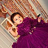 baby, purple_dress, tulle, flower_details, curly_hair, face, celebration, party_decor, balloons, streamers, indoor, child, cute, portrait, festive, smiling, hand_raised, eyes, dress, adorable