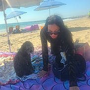 Bella is registered to the contest to win money with this photo: woman, dog, beach, umbrella, ocean, sand, blanket, sunglasses, sunlight, summer, relaxation, curly_hair, pet, vacation, casual, outdoor, shade, bag, waves, sky