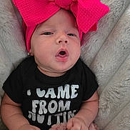 Emmeline is registered to the contest to win money with this photo: baby, infant, headband, pink_bow, black_onesie, text_on_clothing, blanket, soft_texture, cute, expression, face, hands, lying_down, child, adorable, cozy, indoors, newborn, skin, clothing