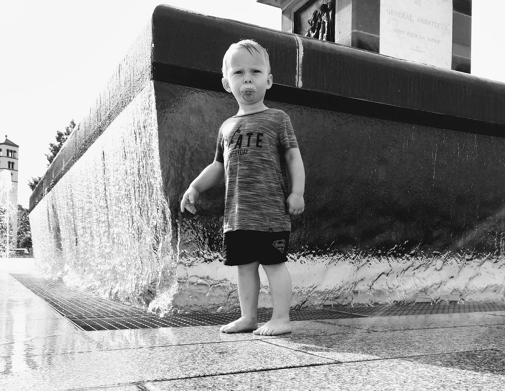 Nolhan participe au concours pour gagner de l'argent avec cette photo : asphalt, black_and_white, city, daytime, flash_photography, fun, grass, happy, human_leg, leisure, monochrome_photography, person, public_space, shorts, standing, style, summer, t_shirt, tints_and_shades, toddler