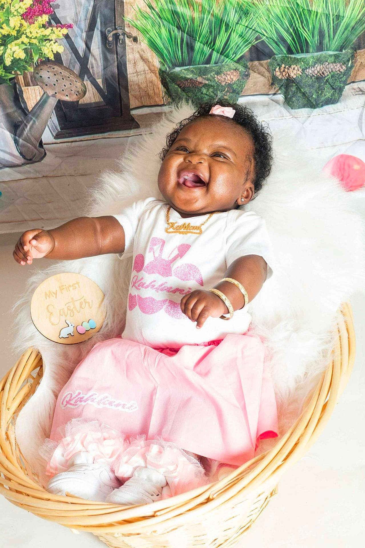 Kahlani is registered to the contest to win money with this photo: baby, girl, smiling, laughing, basket, pink_skirt, white_shirt, bracelets, necklace, bow, fluffy_blanket, wooden_sign, easter, flowers, watering_can, plants, indoors, cute, happy, portrait