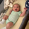 baby, infant, smiling, onesie, blanket, playpen, sock, happy, lying_down, cute, child, infant_clothing, indoor, soft_texture, mattress, newborn, kicking, face, person, cozy