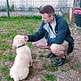 man, dog, puppy, grass, fence, potted_plant, flower, outdoor, pet, animal, casual_clothing, shorts, jacket, sneakers, crouching, feeding, garden, summer, friendship, nature