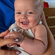 Octavia is registered to the contest to win money with this photo: baby, child, infant, smiling, happy, portrait, face, eyes, mouth, teething, sitting, adult_hand, manicure, nail_polish, gray_clothing, bib, chair, indoor, close_up, family