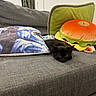 black_cat, kitten, couch, pillow, plush_toy, hamburger_cushion, indoor, gray_fabric, furniture, relaxed, pet, animal, cozy, resting, home, decor, jacket, wall, soft_texture, cute